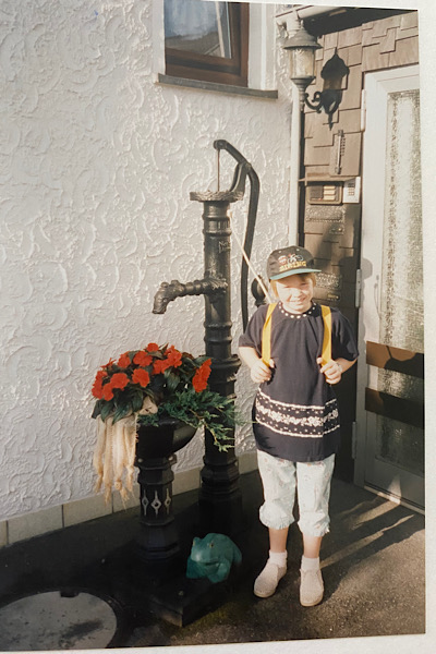 Ein junges Mädchen mit Sonnenkappe steht vor einem Pumpbrunnen und trägt einen Schulranzen